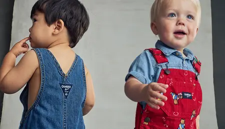 two babies sitting and wearing oshkosh b'gosh overalls